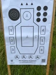 Pistol training target with numbered zones and circular aiming points displayed at an outdoor shooting range.